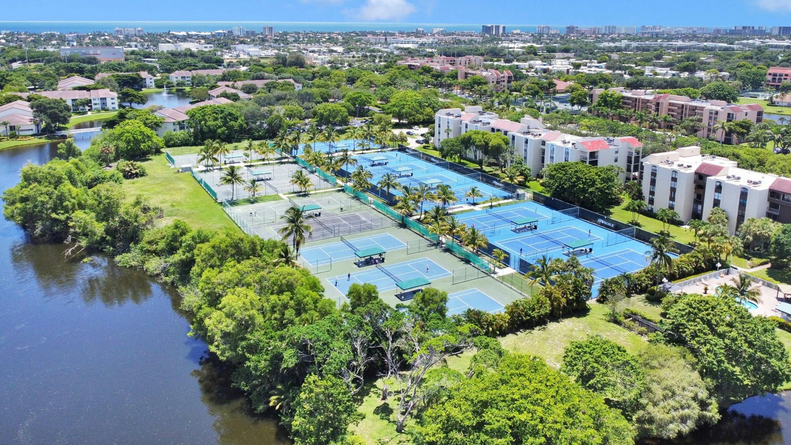 Aerial shot of tennis courts surrounded by lush greenery, apartments, and a river.